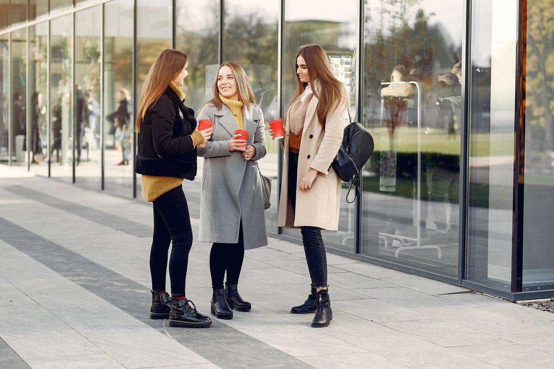 Trzy studentki z kubkami kawy rozmawiają przed budynkiem uczelni
